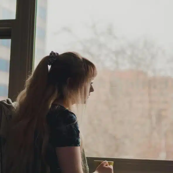 A woman standing by a window looking outside while holding a cup.
