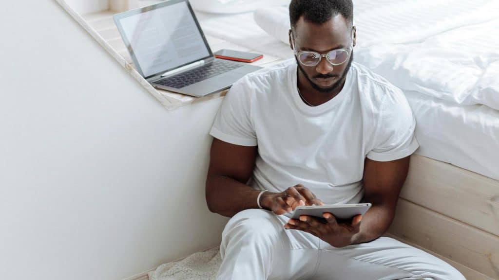 A man sitting by a bed and using a tablet near an open laptop.