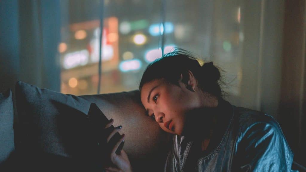 A woman sitting on a couch at night looking at her phone with a thoughtful expression.