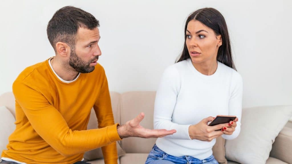 A man in a yellow shirt gestures while speaking to a woman holding a phone.