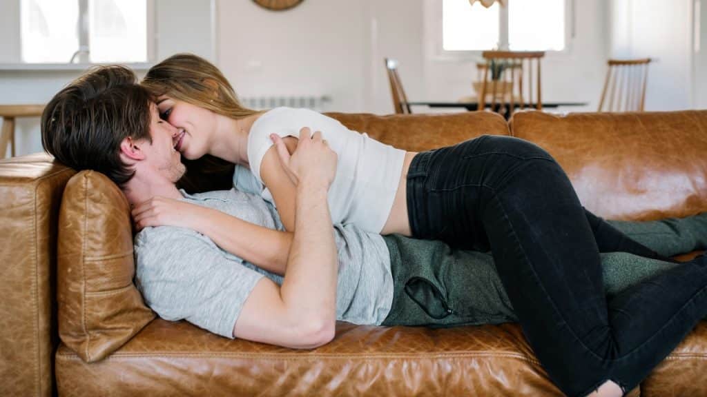 A woman lies on top of a man on a brown leather sofa in a room.