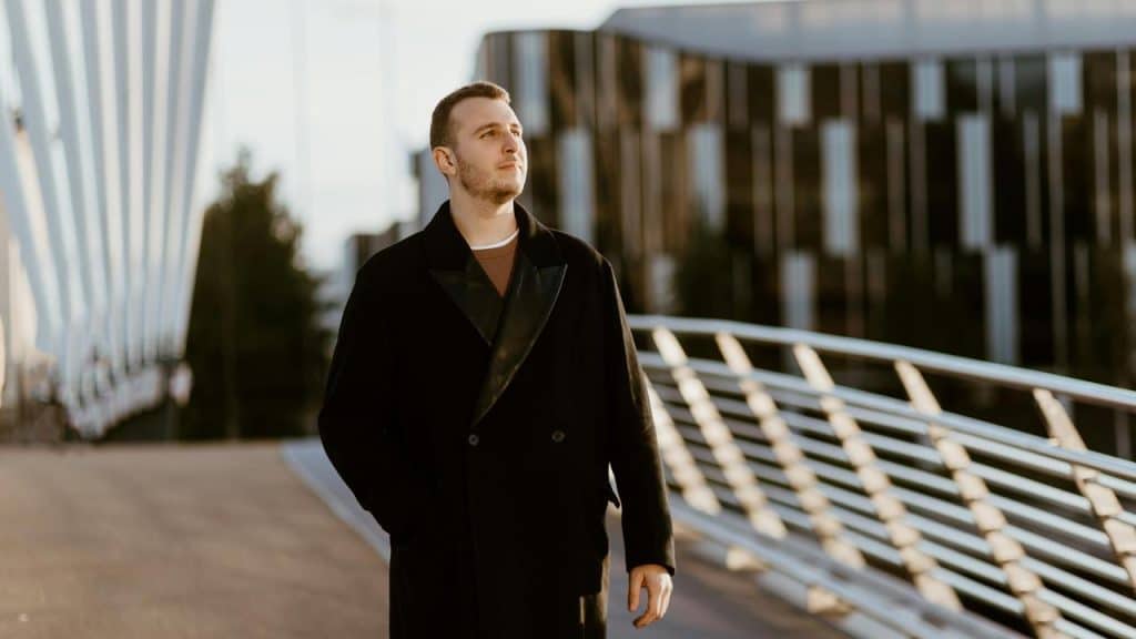 A man in a black coat walking on a modern bridge.