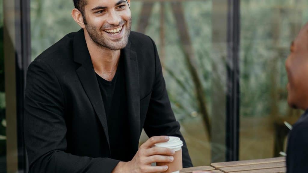 A smiling man in a black blazer holding a coffee cup while chatting at an outdoor café table.