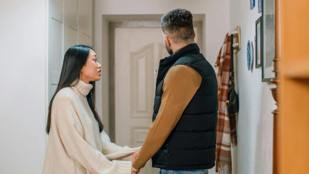 A woman in a turtleneck holds a man's hands while speaking to him indoors.