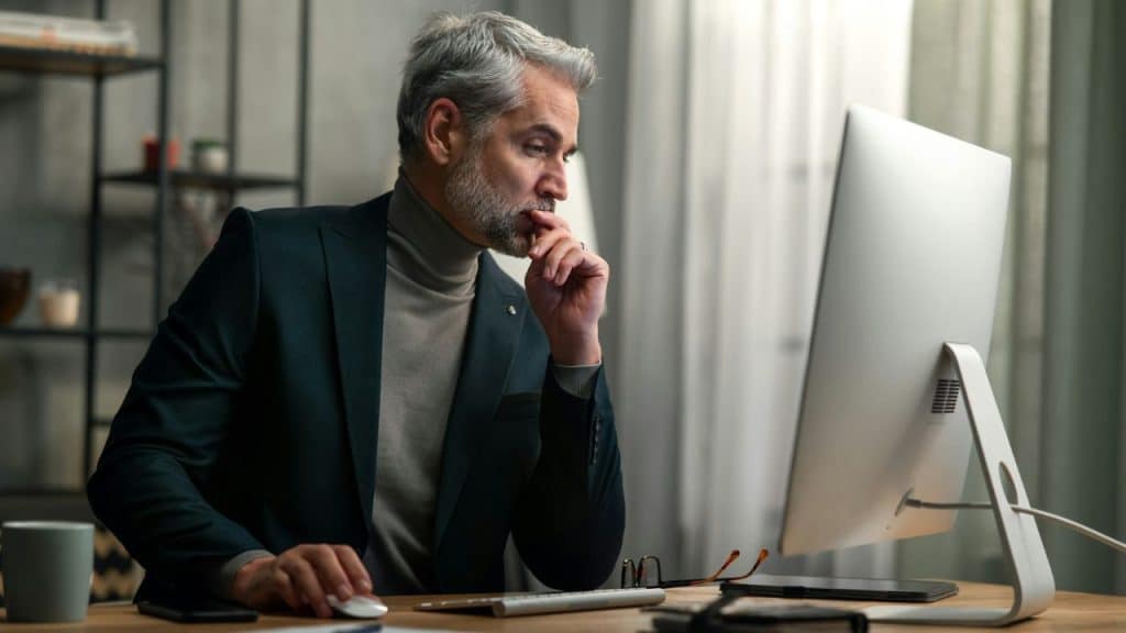 A gray-haired man in a blazer sits at a computer, touching his chin thoughtfully.