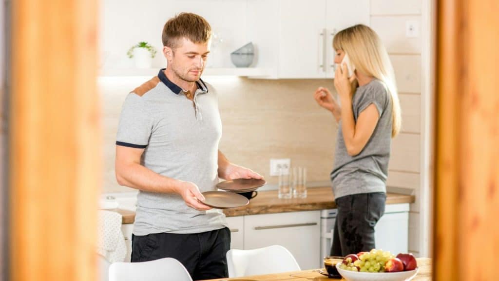A man holding plates stands in a kitchen while a woman talks on the phone.