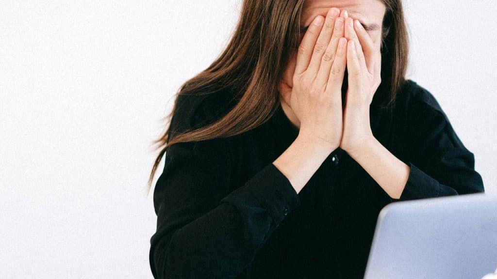 A woman covering her face with her hands while sitting in front of a laptop.