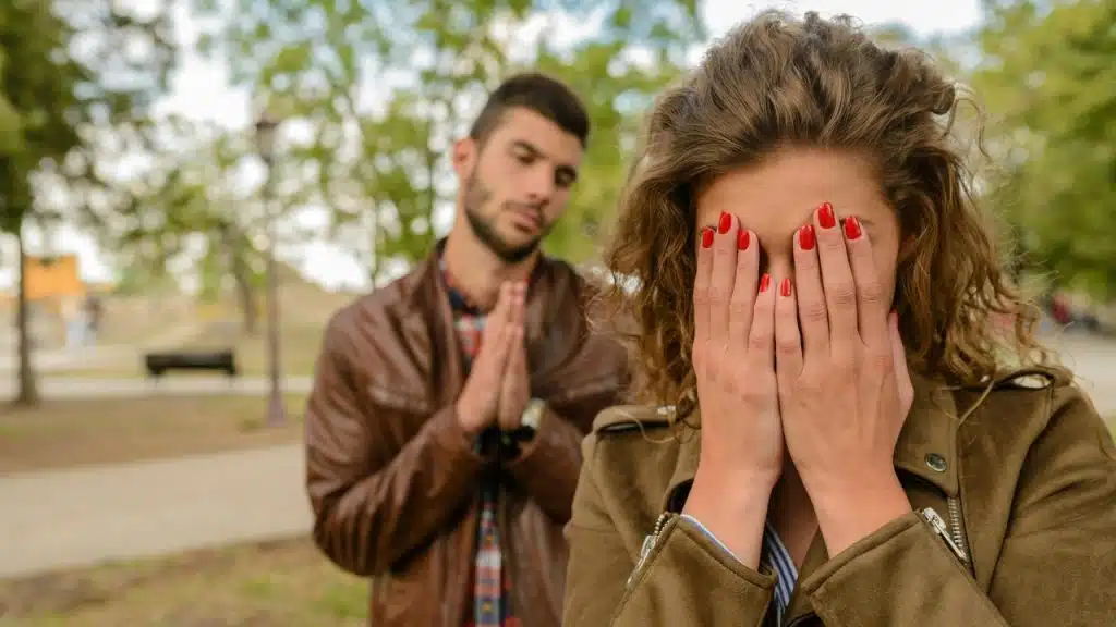 A woman covers her face with her hands while a man stands behind her praying.