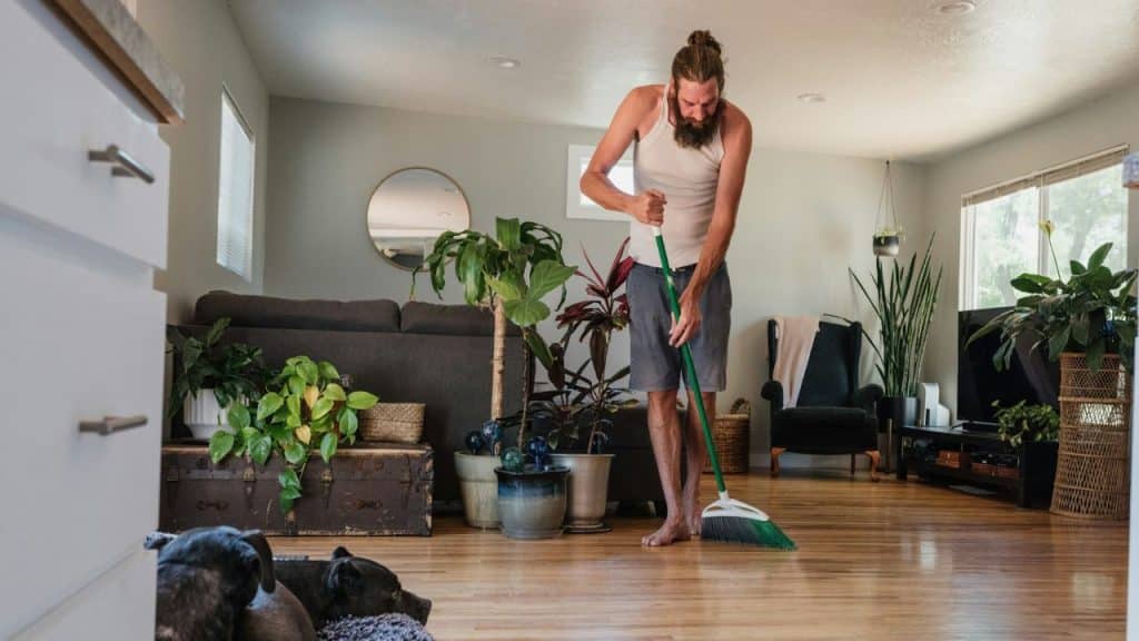 A bearded man with long hair sweeps a wooden floor near several house plants.