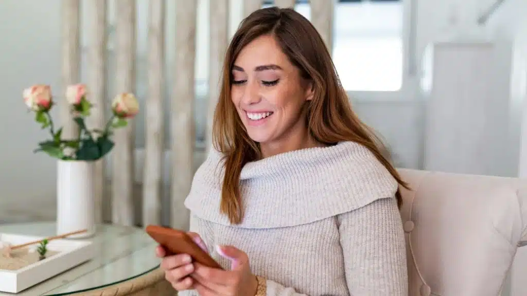 A smiling woman sits in a chair while looking down at a mobile phone.