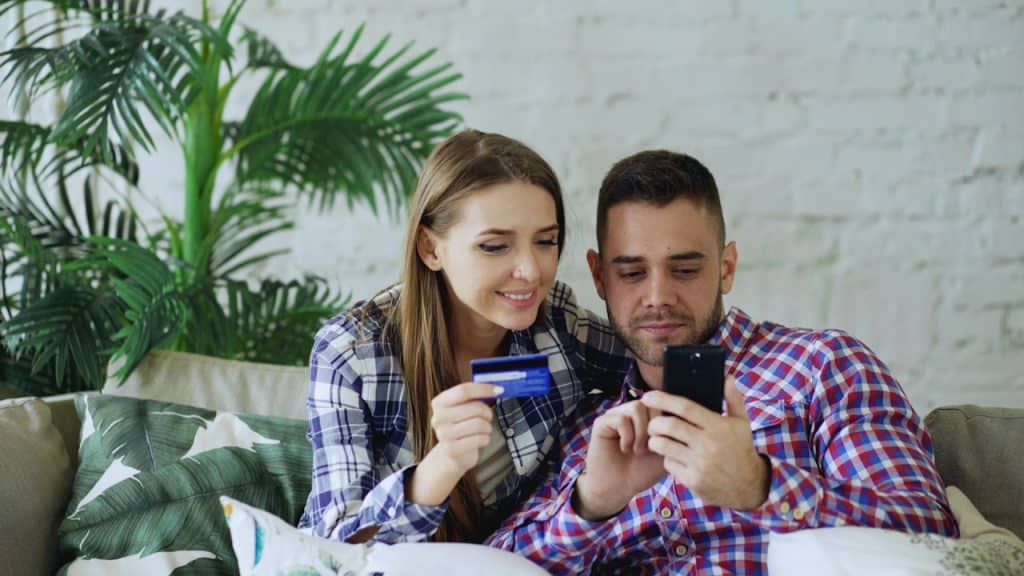 A man and woman sitting on a sofa looking at a smartphone, with the woman holding a blue credit card