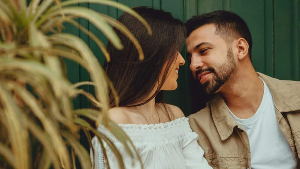 A man and woman look at each other closely while sitting outdoors near green plants.