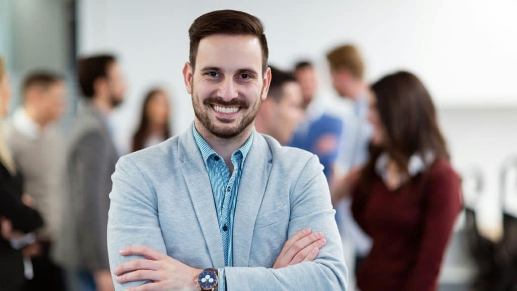 A smiling man in a light blazer stands with arms crossed before a blurred group.
