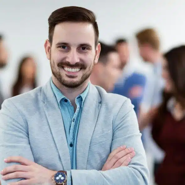 A smiling man in a light blazer stands with arms crossed before a blurred group.