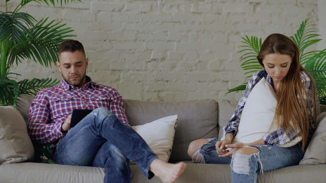 A couple is sitting separated on a sofa, both looking down and engrossed in their own activities.