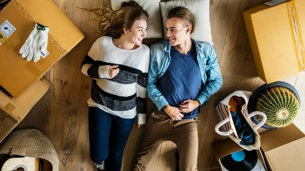 A couple lying on the floor among moving boxes and smiling at each other.