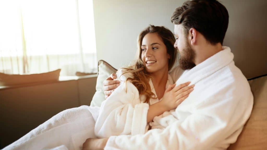 A smiling woman in a white robe rests her head against a man’s shoulder.