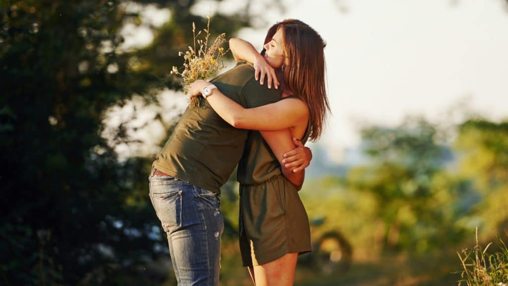 A woman hugs a man outdoors while holding a small bouquet of wildflowers.