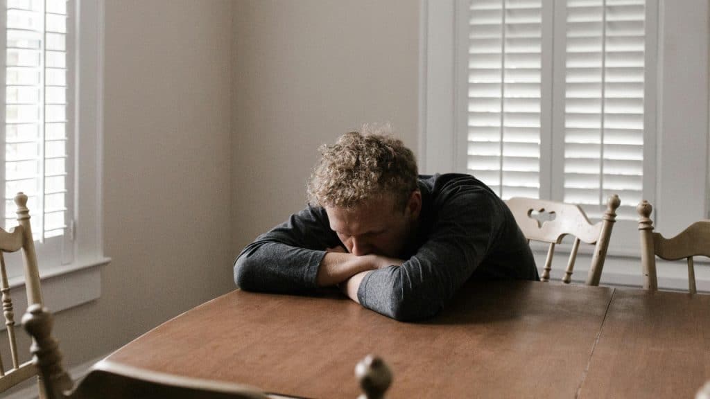 A man rests his head on his crossed arms on a dark wooden table.