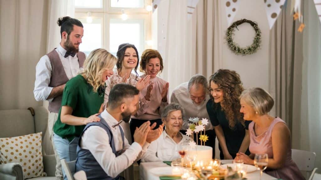 A group of people clap and smile around an elderly woman sitting before a cake.