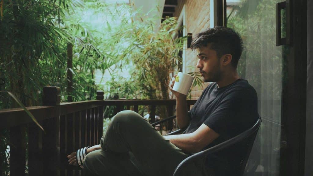 A man sits on a balcony chair drinking from a white mug surrounded by plants.