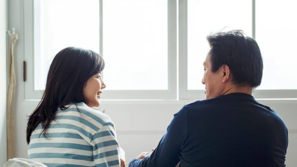 A man and woman sitting with their backs to a large, bright window, looking at and smiling at each other.