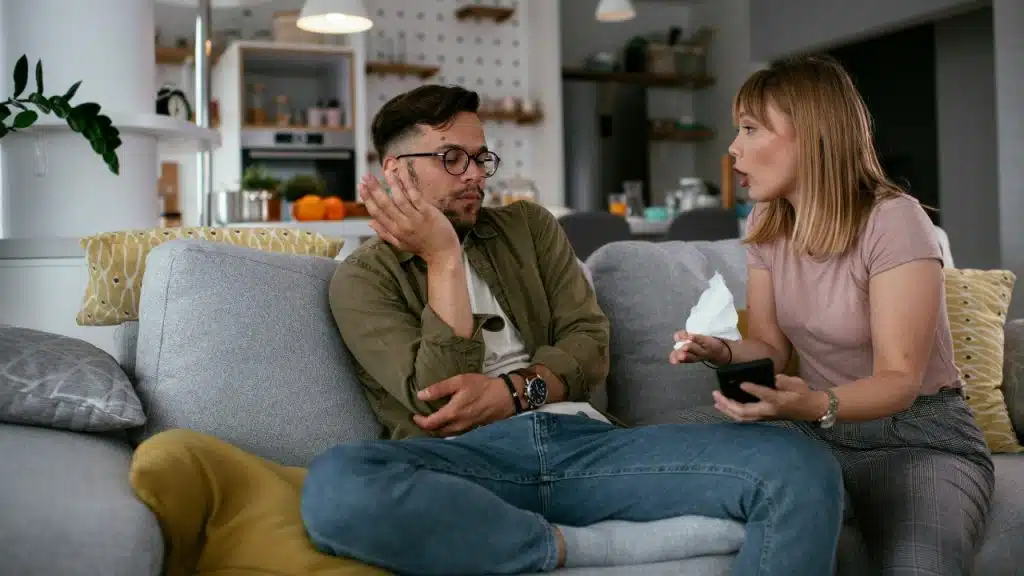 A woman speaks to a man on a couch while holding a phone and tissue.