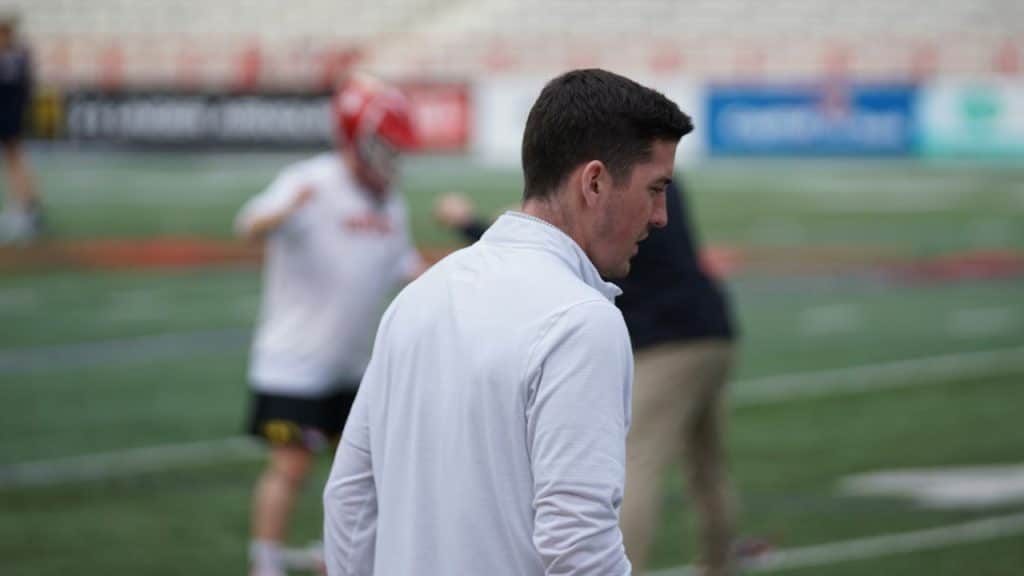A man in a white pullover stands in the foreground of a sports field.