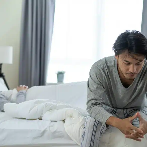 An upset man sitting on the edge of a bed while a woman lies in the bed silently in the background.