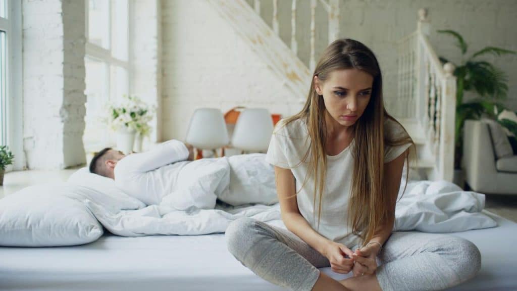 A young woman with a distressed expression sits cross-legged on a bed, while a man lies asleep under the covers in the background.