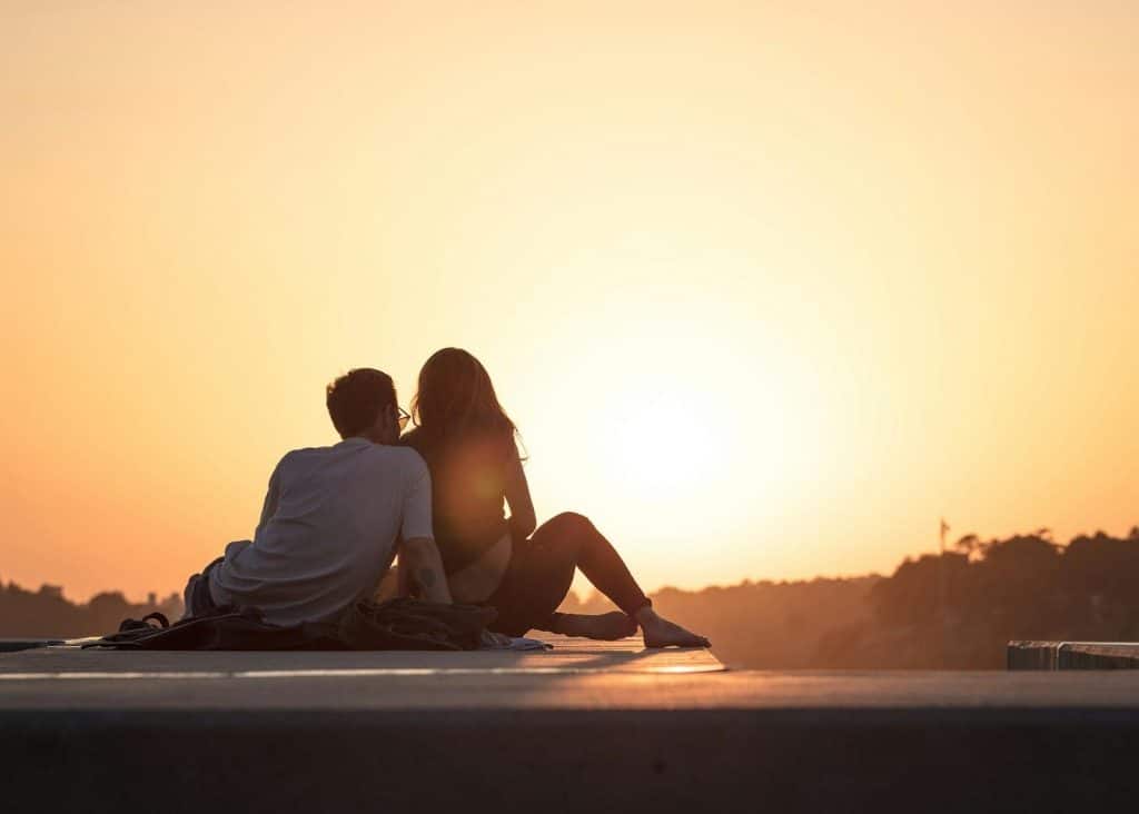 A couple sitting on a ledge watching the sunset together.