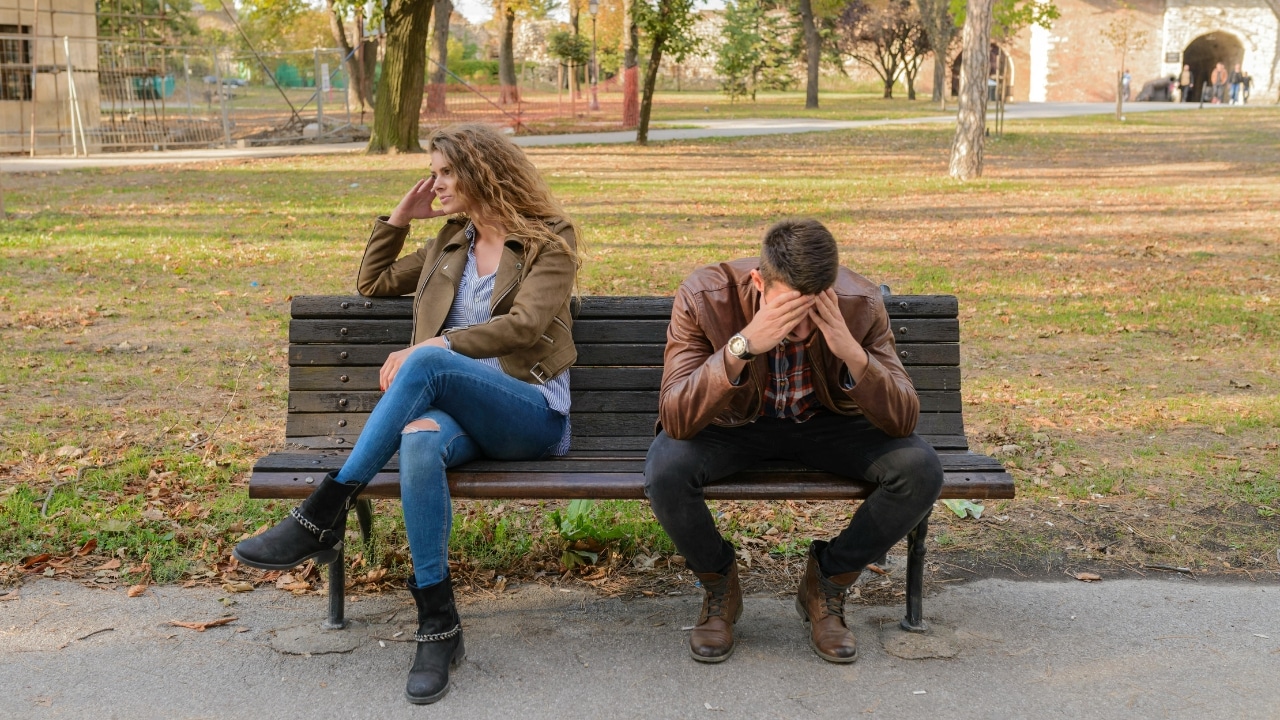 A woman and man sit on a park bench facing away from each other with heads down.