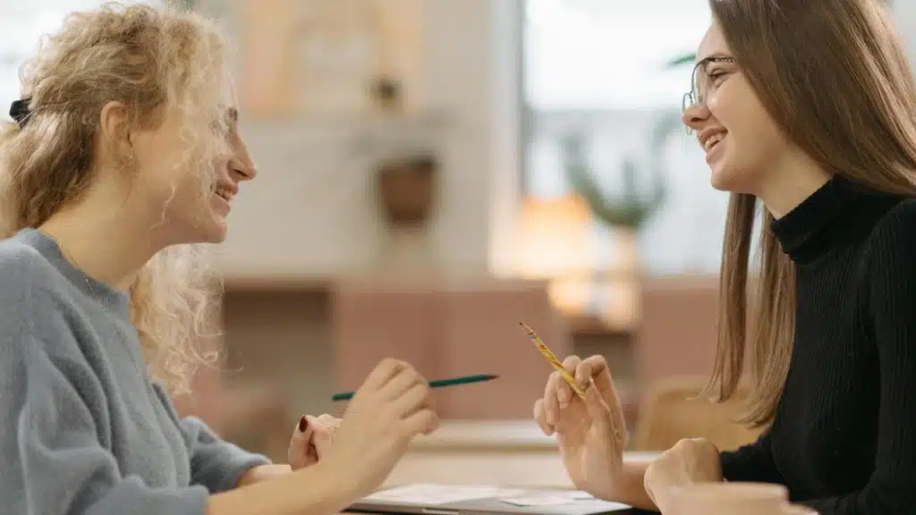 A pair of women smiling and talking while working together at a table.