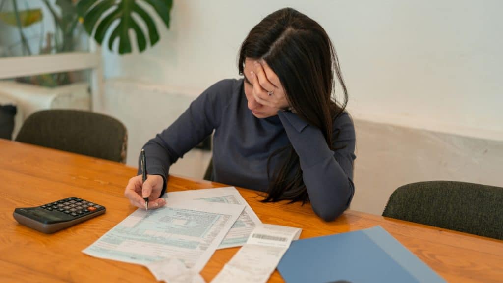 Woman sitting at a wooden table with head in hand, writing on documents near a calculator.
