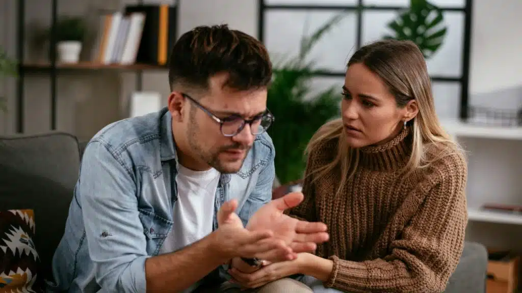A woman in a brown sweater looks at a man in glasses gesturing with his hands.