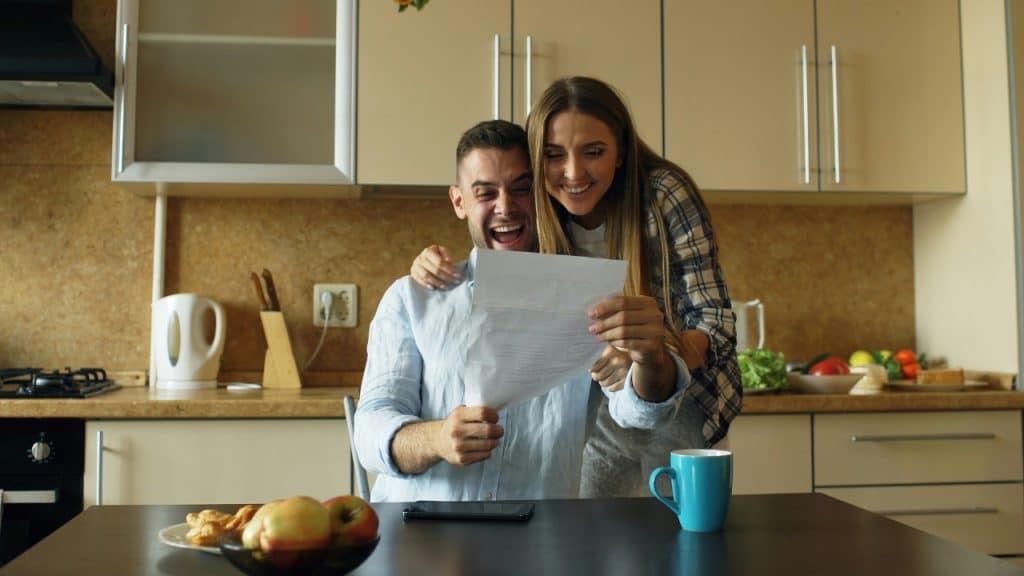 A man and woman stand in a kitchen smiling while looking at a white paper.
