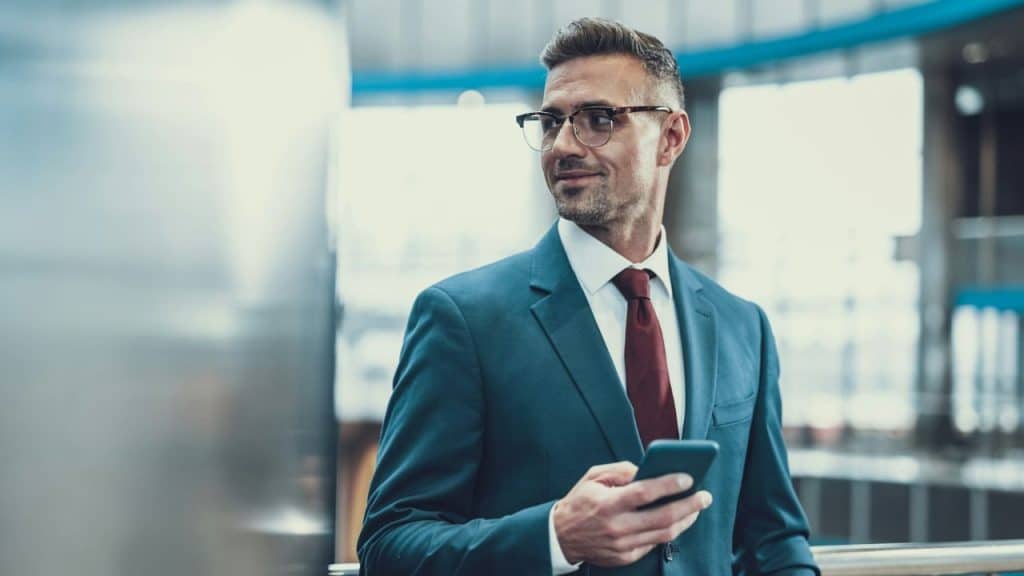 A man in a blue suit and red tie holds a smartphone while looking sideways.