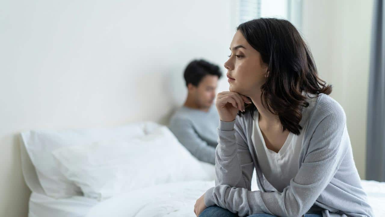 A young woman sits on the edge of a bed while a man sits away from her in the background.
