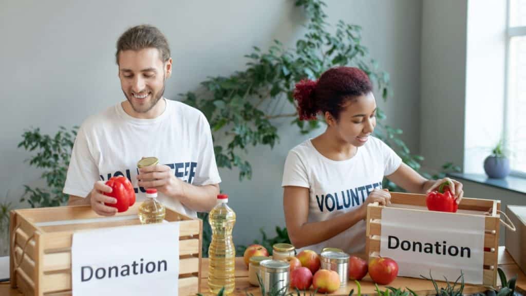 Two people wearing volunteer shirts place fresh produce and canned goods into wooden crates labeled for donation.