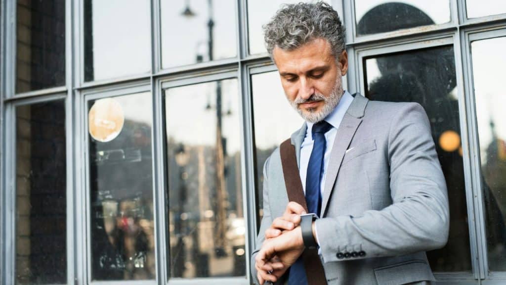 A man in a grey suit looks down at his watch while standing outside a building.