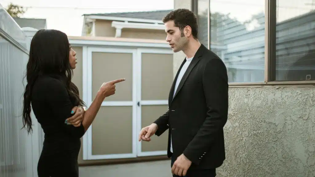 A woman in a black dress points her finger at a man in a black blazer outdoors.