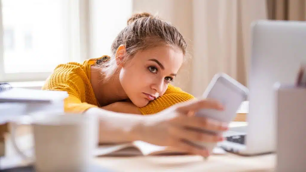 A woman rests her head on a desk while holding a smartphone near a laptop.