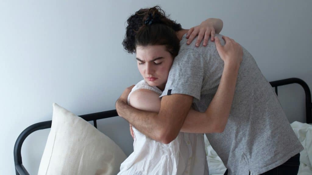 A woman in white and a man in grey hug while sitting on a bed.