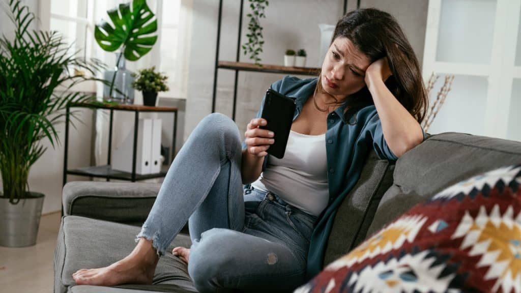 A woman sits on a gray sofa looking down at her phone with a troubled expression.