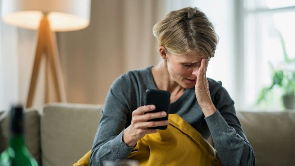 A woman holds a smartphone while resting her forehead against her hand with a distressed expression.
