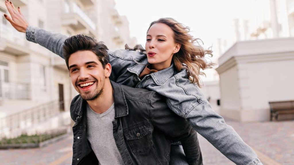 A smiling man giving a piggyback ride to a joyful woman outdoors.