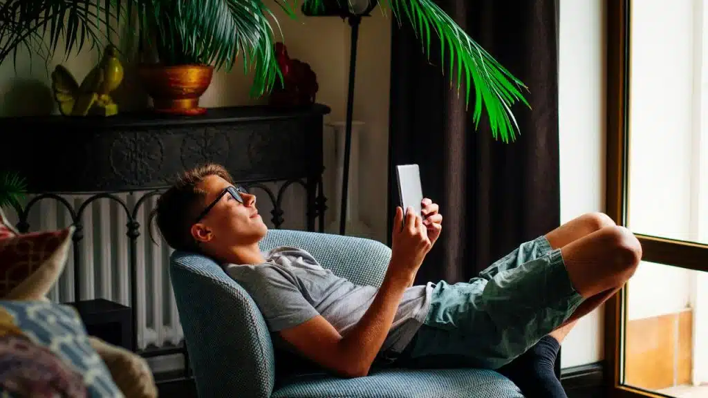 A man lying on a chair reading a tablet near a window.