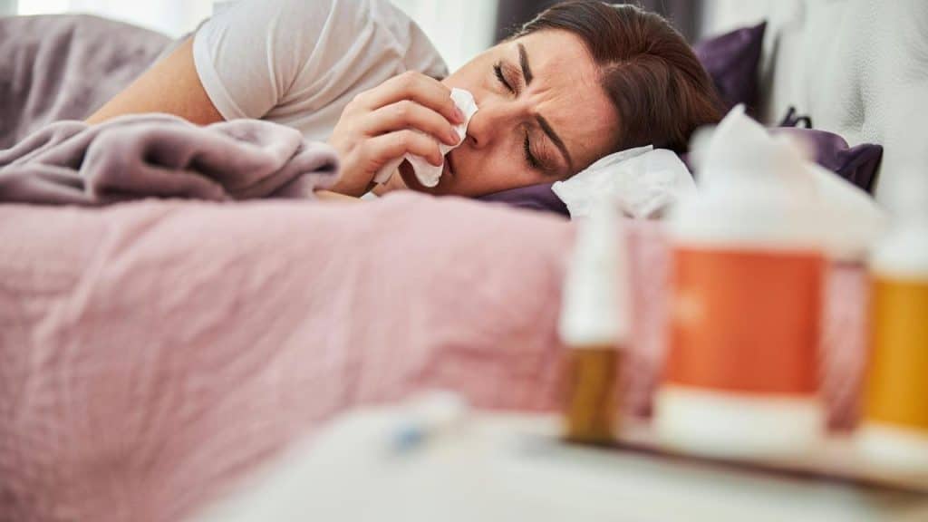 A woman lies in bed holding a tissue, appearing unwell.