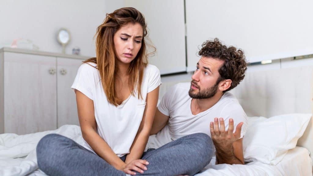 A man and woman sit on a bed while the man gestures with an open hand.