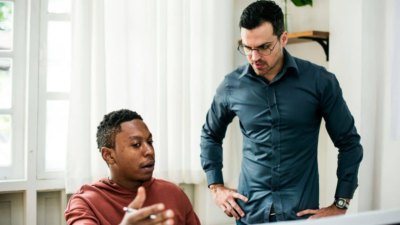 A man in a blue shirt stands over another man who is speaking and gesturing.